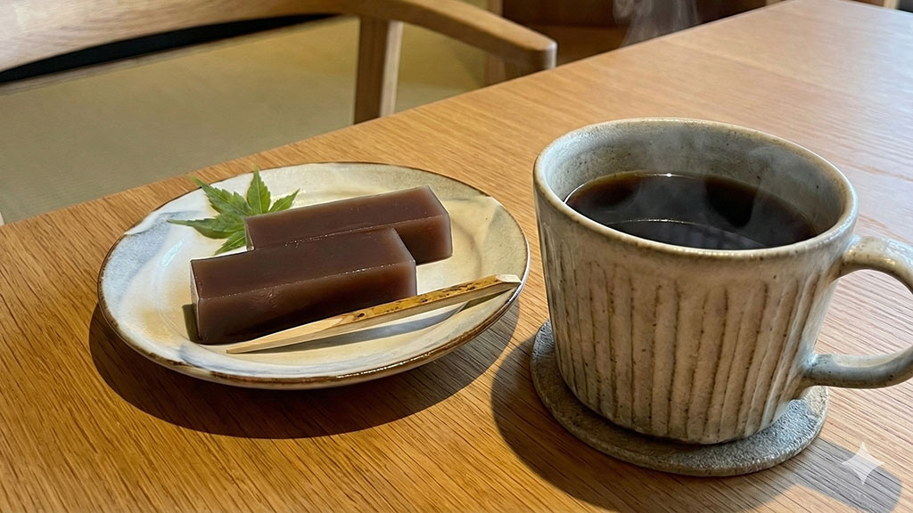 モダンな和カフェの雰囲気で楽しむ水ようかん。上品な小皿に取り分けられた水ようかんの横に、湯気の立つホットブラックコーヒーのカップが置かれ、和菓子とコーヒーのペアリングを提案している一枚。